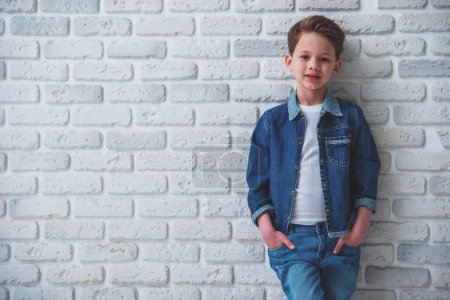Foto per Elegante bambino in abiti jeans sta guardando la fotocamera e sorridente, in piedi con le mani in tasca contro muro di mattoni bianchi - Immagine Royalty Free