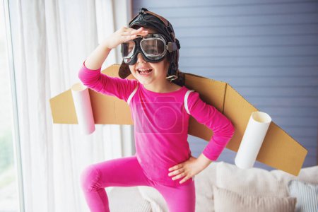 Foto de Linda niña vestida como un piloto con alas de juguete sonríe y escanea el horizonte mientras juega en casa - Imagen libre de derechos