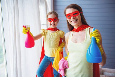 Téléchargez les photos : Belle mère et sa jolie petite fille habillée comme des super-héros tiennent pulvérisateur, regardant la caméra et souriant tout en nettoyant la maison - en image libre de droit