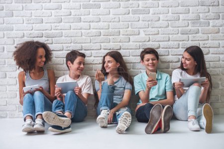 Grupo de chicos y chicas adolescentes está utilizando gadgets, hablando y sonriendo, sentado contra la pared de ladrillo blanco

