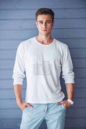 Portrait of handsome young man in casual clothes looking at camera, standing with hands in pockets against gray wall