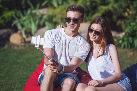 Foto de Joven hermosa pareja en ropa casual y gafas de sol está sonriendo mientras se hace selfie utilizando el teléfono inteligente y el monópodo, sentado en sillas bolsa de frijoles al aire libre - Imagen libre de derechos