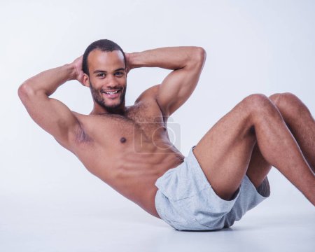 Photo for Handsome sportsman is doing abs, looking at camera and smiling while working out topless, isolated on white background - Royalty Free Image