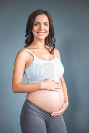 Photo for Beautiful pregnant young woman in casual clothes is touching her bare tummy, looking at camera and smiling, on a gray background - Royalty Free Image