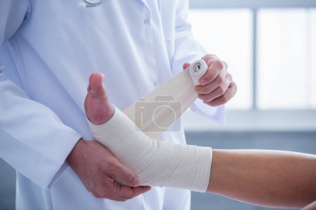 geschnittenes Bild eines gut aussehenden Arztes, der das verletzte Bein einer Frau bei der Arbeit in seinem Büro bandagiert
