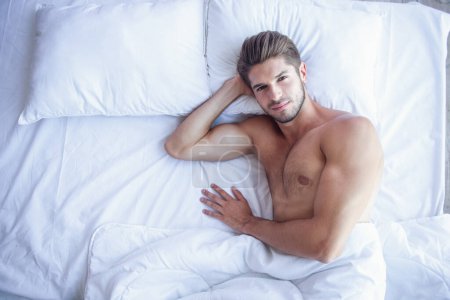 Téléchargez les photos : Vue du dessus du jeune homme musclé sexy regardant la caméra avec un sourire sensuel tout en étant couché au lit - en image libre de droit