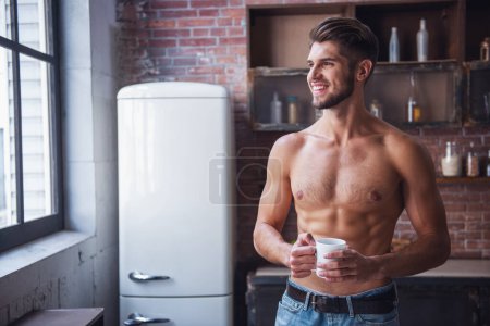 Photo for Sexy young man with bare torso is holding a cup, looking out the window and smiling while standing in kitchen at home - Royalty Free Image