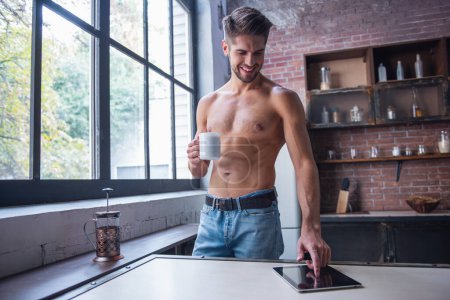 Photo for Sexy young man with bare torso is holding a cup, using a digital tablet and smiling while standing in kitchen at home - Royalty Free Image