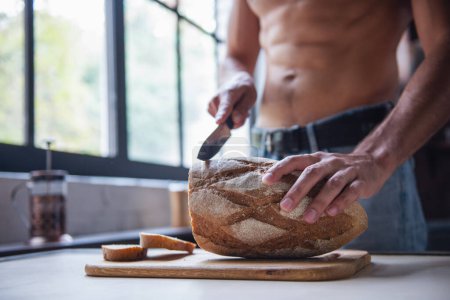Photo for Cropped image of sexy young man with bare torso cutting bread while standing in kitchen at home - Royalty Free Image
