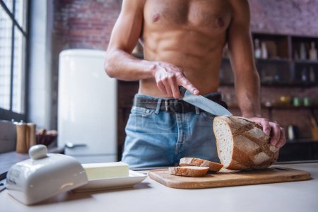 Photo for Cropped image of sexy young man with bare torso cutting bread while standing in kitchen at home - Royalty Free Image
