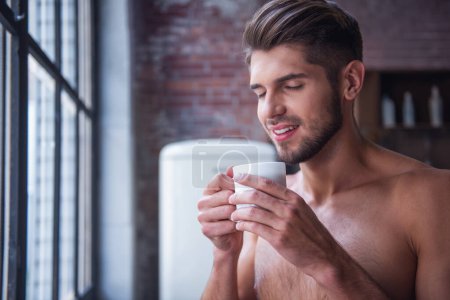 Photo for Sexy young man with bare torso is holding a cup, smelling it and smiling while standing in kitchen at home - Royalty Free Image