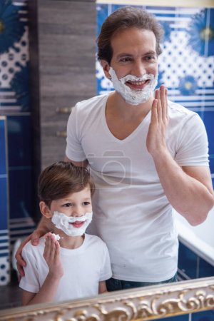 Téléchargez les photos : Père et fils appliquent de la mousse à raser sur leur visage et sourient tout en se rasant dans la salle de bain - en image libre de droit