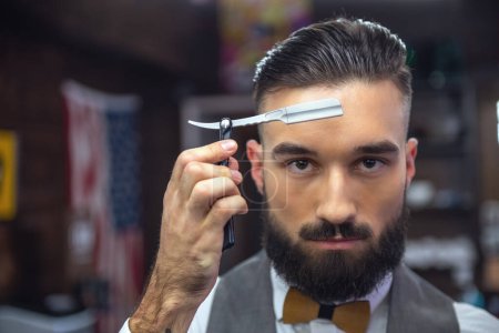 Foto de Guapo peluquero barbudo en ropa clásica con estilo es la celebración de una navaja de afeitar recta mientras está de pie en la barbería - Imagen libre de derechos