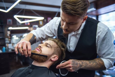 Foto de Hombre barbudo guapo está recibiendo el peinado de barba por peluquería en la barbería - Imagen libre de derechos