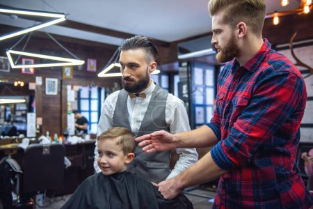Téléchargez les photos : Mignon petit garçon visite le salon de coiffure. Son père et le coiffeur discutent de la coiffure du garçon. - en image libre de droit