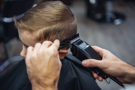 Téléchargez les photos : Vue latérale de mignon petit garçon se faire couper les cheveux par coiffeur au salon de coiffure - en image libre de droit