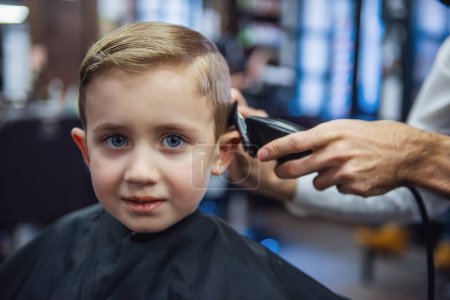 Téléchargez les photos : Mignon petit garçon regarde la caméra tout en se faisant couper les cheveux par un coiffeur au salon de coiffure - en image libre de droit