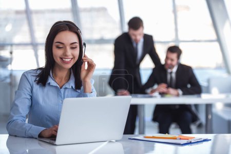 Foto de Atractiva mujer de negocios en auriculares está sonriendo mientras trabaja con el ordenador portátil en la oficina, sus colegas en el fondo - Imagen libre de derechos
