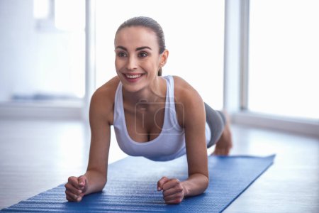 Photo for Beautiful girl in sportswear is smiling while doing plank exercise on yoga mat - Royalty Free Image