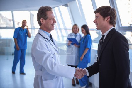 Téléchargez les photos : Beau médecin mature et jeune homme d'affaires serrent la main et sourient tout en se tenant dans le hall de l'hôpital - en image libre de droit