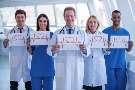 Téléchargez les photos : Une équipe réussie de médecins tient des graphiques de la fréquence cardiaque, regarde la caméra et sourit tout en restant debout dans la salle d'hôpital - en image libre de droit