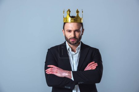 Foto de Hombre de negocios guapo en traje y con una corona en la cabeza está mirando a la cámara mientras está de pie con los brazos cruzados sobre fondo gris - Imagen libre de derechos