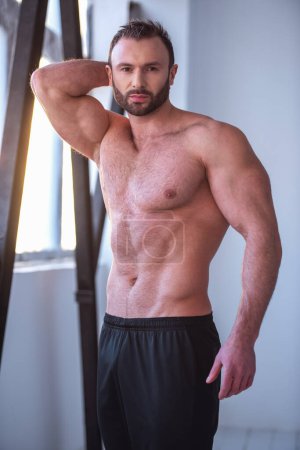 Foto de Retrato de un hombre musculoso guapo con el torso desnudo mirando a la cámara mientras está de pie en el gimnasio - Imagen libre de derechos