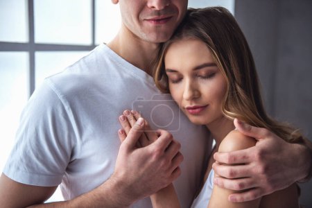 Photo for Beautiful young couple is hugging and smiling while standing near the window at home - Royalty Free Image