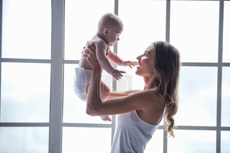 Photo for Beautiful young mom is playing with her cute baby and smiling while standing near the window at home - Royalty Free Image