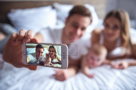 Photo for Beautiful young parents are spending time with their baby. Dad is doing selfie using a smartphone - Royalty Free Image