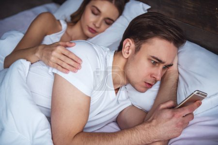 Foto de Una pareja joven tiene un problema. Chico está charlando con un teléfono inteligente mientras su novia está durmiendo y abrazándolo - Imagen libre de derechos
