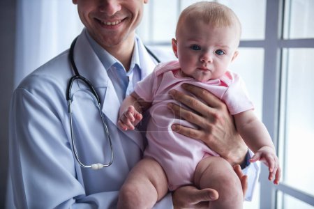 Photo for Handsome young doctor is holding a cute baby and smiling - Royalty Free Image