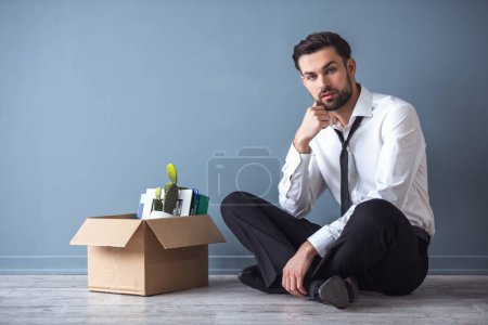 Foto de Que te despidan. Hombre de negocios guapo en ropa formal está sentado en el suelo cerca de la caja con sus cosas, sobre fondo gris - Imagen libre de derechos