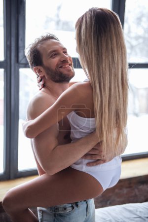 Photo for Beautiful couple is smiling and hugging while spending time together at home. Man is holding his girlfriend in arms - Royalty Free Image