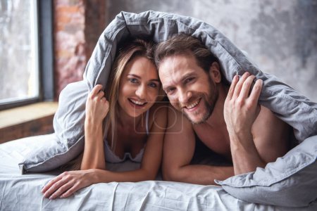 Téléchargez les photos : Beau couple regarde la caméra et souriant tout en passant du temps ensemble dans le lit à la maison - en image libre de droit