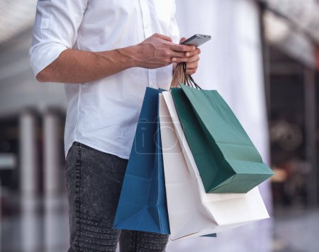 Foto de Imagen recortada de chico guapo con bolsas de compras usando un teléfono móvil mientras hace compras en el centro comercial - Imagen libre de derechos