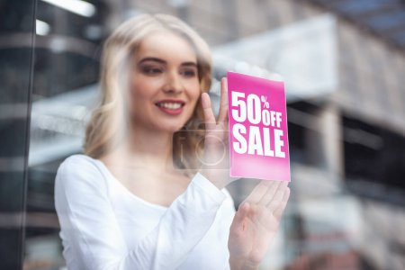 Téléchargez les photos : Belle assistante de vente met sur la vitrine une annonce sur les ventes et souriant - en image libre de droit