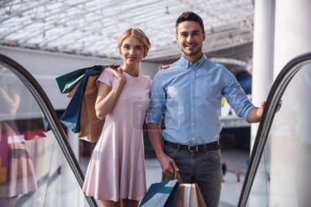 Foto de Hermosa pareja con bolsas de compras está mirando a la cámara y sonriendo mientras está de pie en la escalera mecánica en el centro comercial - Imagen libre de derechos