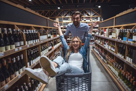 Photo for Beautiful couple is having fun while doing shopping at the supermarket. Girl is sitting in the shopping cart - Royalty Free Image