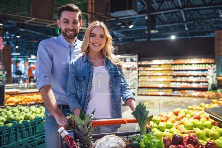 Téléchargez les photos : Beau couple avec un panier regarde la caméra et souriant tout en faisant des achats au supermarché - en image libre de droit