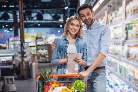 Photo for Beautiful couple is looking at camera and smiling while doing shopping at the supermarket - Royalty Free Image