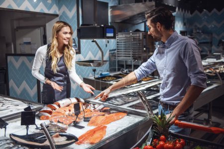 Photo for At the supermarket. Beautiful young female worker is offering fish to the handsome customer and smiling - Royalty Free Image