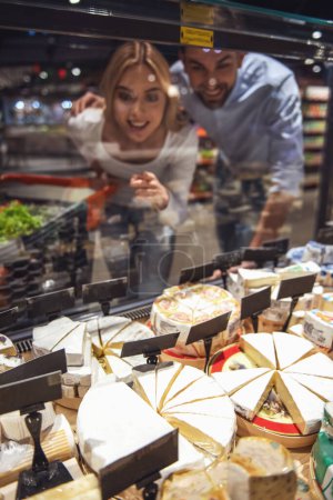 Foto de Hermosa pareja joven está eligiendo queso y sonriendo mientras hace compras en el supermercado - Imagen libre de derechos