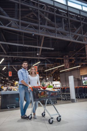 Photo for Beautiful young couple is looking at camera and smiling while doing shopping at the supermarket - Royalty Free Image
