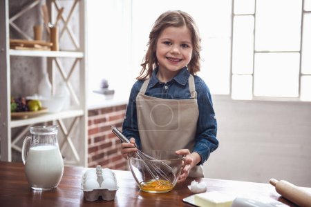 Téléchargez les photos : Petite fille mignonne dans le tablier regarde la caméra et souriant tout en fouettant des œufs pour la cuisson dans la cuisine - en image libre de droit