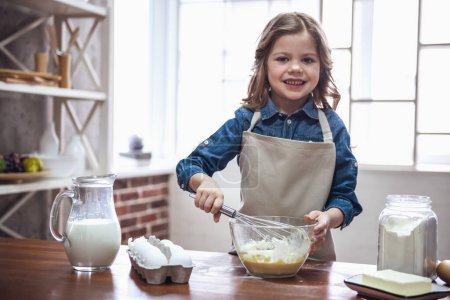 Téléchargez les photos : Petite fille mignonne dans le tablier regarde la caméra et souriant tout en mélangeant la pâte pour la cuisson dans la cuisine - en image libre de droit