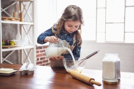Photo for Cute little girl in apron is adding milk to eggs while preparing dough for baking in the kitchen - Royalty Free Image