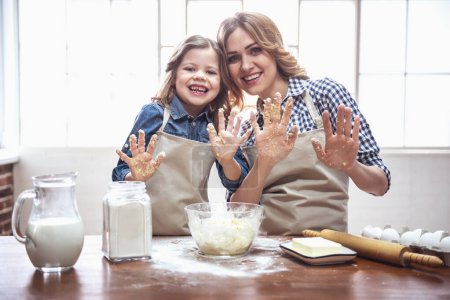 Foto de Linda niña y su hermosa mamá en delantales están mostrando las palmas, mirando a la cámara y sonriendo mientras amasa la masa en la cocina - Imagen libre de derechos