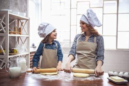 Téléchargez les photos : Petite fille mignonne et sa belle maman dans des tabliers et des chapeaux de chef sourient tout en aplatissant la pâte à l'aide d'un rouleau à pâtisserie dans la cuisine - en image libre de droit