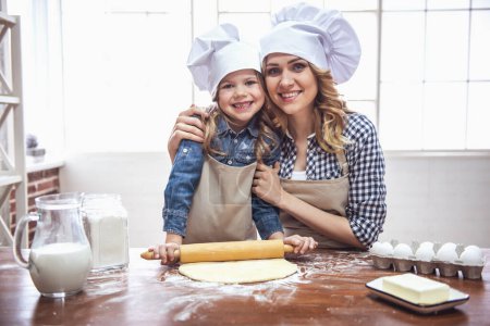 Photo for Cute little girl and her beautiful mom in aprons and chef hats are smiling while flattening the dough using a rolling pin in the kitchen - Royalty Free Image